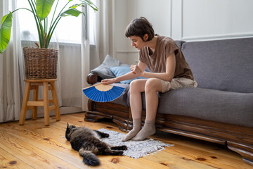 Caring young woman fan overheating cat on floor in hot apartment with no AC. Pet owner struggle in...