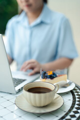 Cup of black coffee and dessert cake on cafe table with blurred woman working on laptop in background, concept of remote work, freelance lifestyle, outdoor study, productivity and modern relaxation.