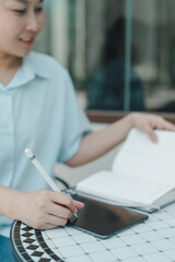 Smiling woman working on creative project using digital tablet and stylus while holding notebook, sitting at outdoor café table, combining handwritten notes and digital tools in casual setting.