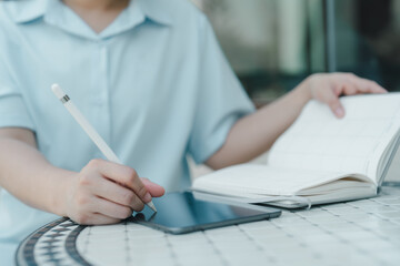 Young woman writing on tablet with stylus and referencing notebook, enjoying remote work or study session in modern outdoor environment, blending traditional and digital productivity tools.