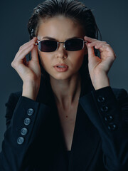Stylish woman wearing black sunglasses and dark blazer poses confidently against a muted gray...