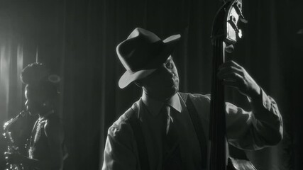 Black and white shot of old fashioned Caucasian man wearing fedora hat and playing upright bass while performing on stage in jazz club, retro vibes