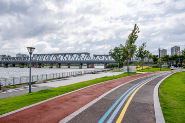 Scenic riverside exercising trail in Ningbo, China, featuring dedicated walking, running, and cycling lanes with a steel railway bridge in the background.A well-maintained fitness route for community.