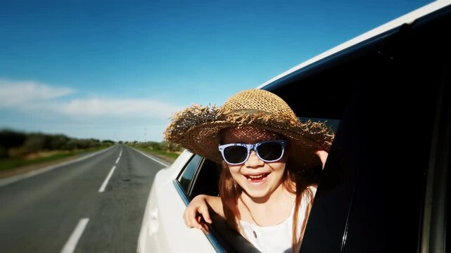 Laughing joyful little girl looking out of the car window riding on a sunny summer day along a country road. Sincere emotions of happiness and carelessness during a family trip to nature