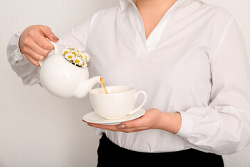 Beautiful young woman pouring chamomile tea from teapot into cup near white wall