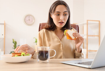 Beautiful young businesswoman eating tasty sandwich at her workplace in office