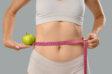 Beautiful young woman with apple measuring her waist on grey background, closeup. Weight loss...