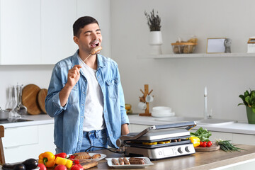 Young man with cooked tasty sausages and vegetables on modern electric grill at table in kitchen