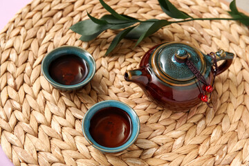 Wicker mat with teapot, cups of tea and leaves on purple background, closeup
