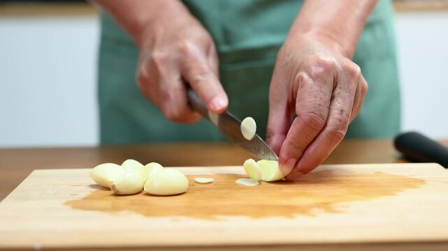 Closeup of hands slicing fresh garlic on a wooden cutting board in kitchen. Perfect for cooking, food prep, healthy eating, herbs
