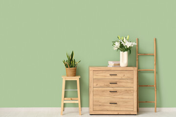 Vase with lily flowers on wooden chest of drawers near green wall in room