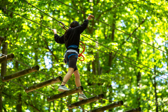 Fototapeta Child with harness crossing a rope bridge in a fun adventure park for kids. Outdoor forest activity focused on balance, courage, and safety. Adventure park with kids sport activities.