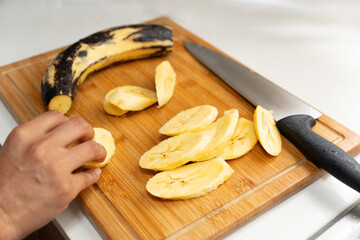 Photograph of plantains cut on a wooden chopping board with a knife, with one hand placing the...