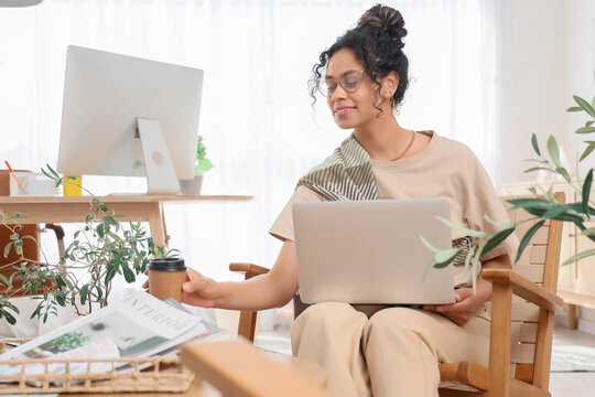 Young African-American graphic designer with laptop taking coffee cup from table in office