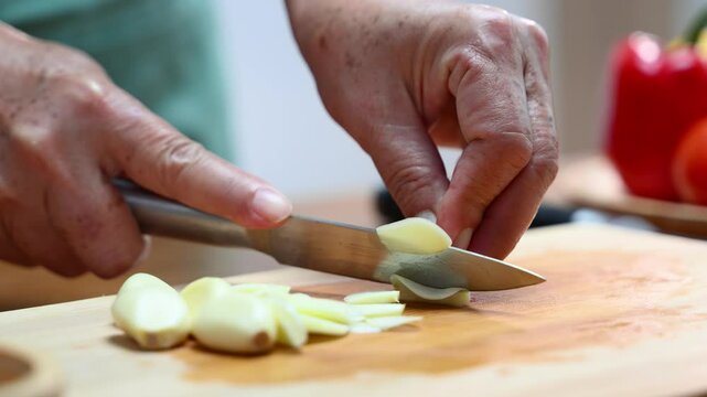 Closeup of hands slicing fresh garlic on a wooden cutting board in kitchen. Perfect for cooking, food prep, healthy eating, herbs