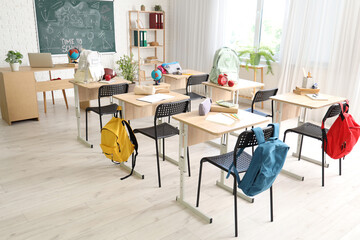 School desks with backpacks and blackboard in light classroom