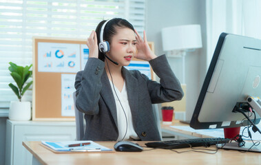 An Asian woman sits at her desk in a call center, wearing a headset. Her expression reveals stress,...