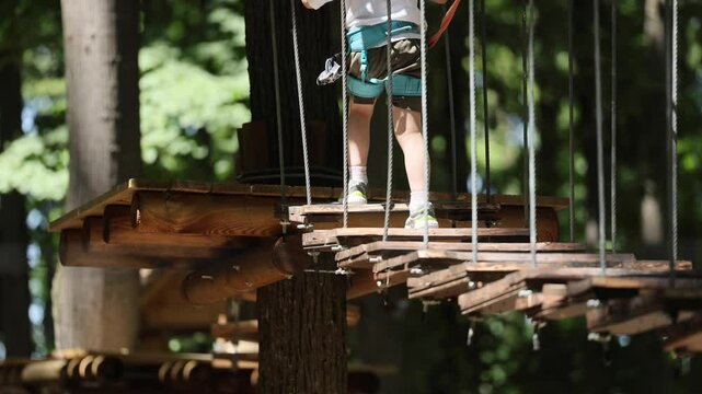 Adventure park 4k video. Child with harness crossing a rope bridge in a fun adventure park for kids. Outdoor forest activity focused on balance, courage, and safety. kids sport activities video.