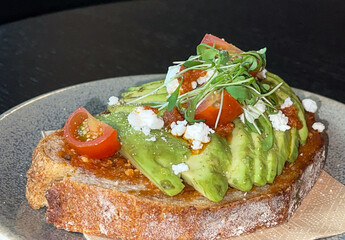 Avocado toast bread, slices garnished with tomatoes and feta cheese, microgreens and sauce, in ceramic plate on black wood table, high angle view, copy space