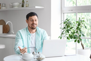 Young male teacher with laptop and notebook giving online lesson at home