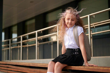 young girl with backpack sitting on school bench in outdoor setting. blonde hair blowing in breeze,...