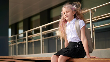 young girl smiling with backpack sits outside school, wind in hair. wearing uniform, ready for new academic year, embodying curiosity and anticipation for education journey