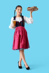 Beautiful young happy Asian Octoberfest waitress with snacks and glass of beer on blue background