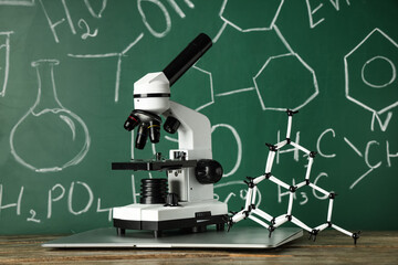 Modern microscope, laptop and molecular model on wooden table near chalkboard with chemical formulas