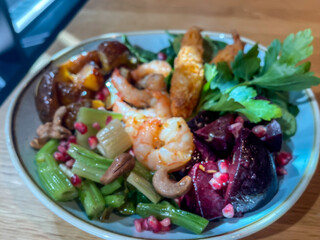 Shrimps and fish sticks on colorful vegetable bed in blue ceramic plate on wood table, sunray highlights, red beets, stalk celery, nut and pomegranate garnish, spinach, top down view