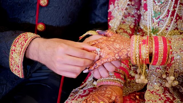 Groom is putting on the ENGAGEMENT ring on Traditional henna painted bridal hand. Red color is the traditional Indian clothes, Elements of hindu wedding. 