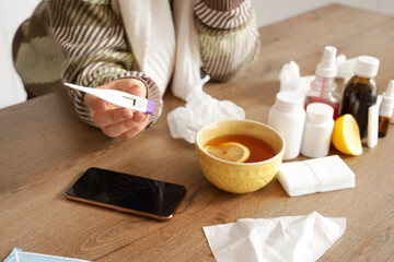 Sick woman with thermometer and flu medications on table in kitchen, closeup