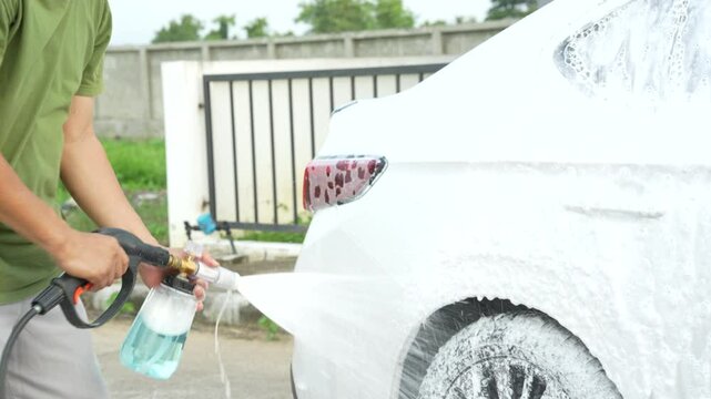 A middle-aged Asian man carefully washes his car using water and foam. Each step from high-pressure rinsing to scrubbing and drying ensures the vehicle is spotless, well-maintained, and presentable