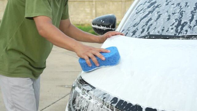 A middle-aged Asian man carefully washes his car using water and foam. Each step from high-pressure rinsing to scrubbing and drying ensures the vehicle is spotless, well-maintained, and presentable