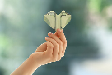 Hand holding dollar banknote in shape of heart on blurred background. Donation concept