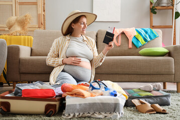 Young pregnant woman with passport, clothes, travelling accessories and full suitcase at home