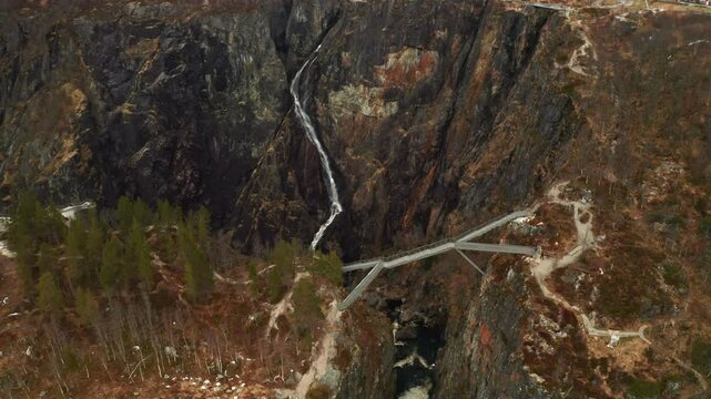 Voringsfossen waterfall cascades down rugged dark rock face with modern bridge crossing over canyon. Dramatic scale of natural wonder in Norway