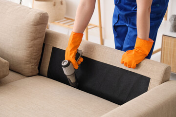 Male janitor with modern handheld vacuum cleaner cleaning sofa in living room