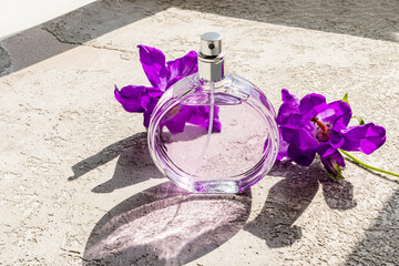 A beautiful bottle of women's perfumes stands on a concrete surface with lilac iris flowers in the sun. A blank bottle mockup. Product presentation.