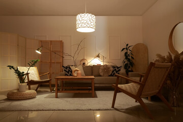 Interior of stylish living room with cozy grey sofa, armchairs, coffee table and glowing lamps at...