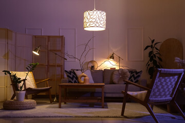 Interior of stylish living room with cozy grey sofa, armchairs, coffee table and glowing lamps at night
