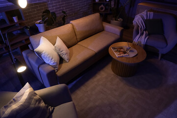 Interior of stylish living room with cozy sofa, coffee table, armchairs and glowing lamps at night