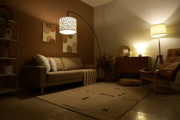 Interior of stylish living room with cozy sofa, armchair and glowing lamps at evening