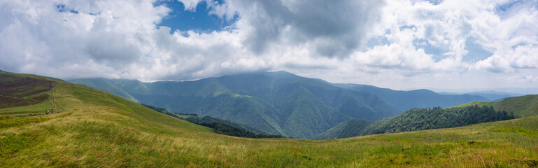 A panoramic view of rolling green hills and dense forests under a dramatic, cloudy sky. The layered landscape stretches into the distance, capturing the raw beauty of a mountainous countryside