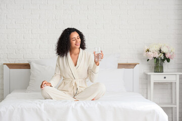 Young African-American woman with glass of water sitting in bedroom