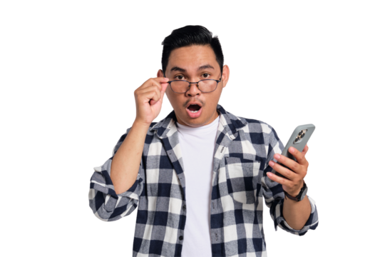 Surprised young Asian man in casual shirt holding smartphone, taking off his glasses, looking at camera with amazed face isolated on transparent background