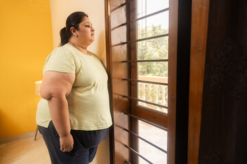 Obese indian woman feels lonely at home. Plus size lady standing alone near window . Stress and...