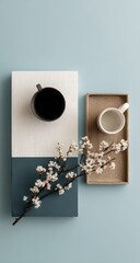 Two trays, one dark and one light, hold mugs and a blossoming branch on a pale blue background; minimalist, serene aesthetic