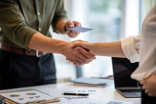Two business professionals shaking hands over a table with financial reports and a laptop symbolizing partnership and agreement - Powered by Adobe