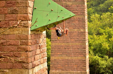 Horizontal photo capturing a woman hanging from a green overhanging climbing wall outdoors. Action...