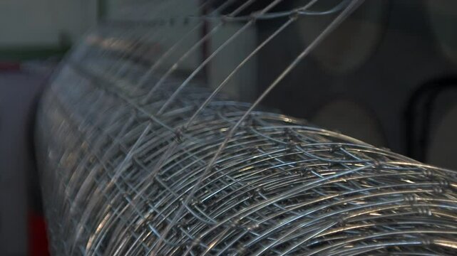 A close-up view of a large roll of galvanized wire mesh, likely a fixed-knot fence, in an industrial setting. The metallic silver wires are woven together to form a grid pattern, and the roll is posit